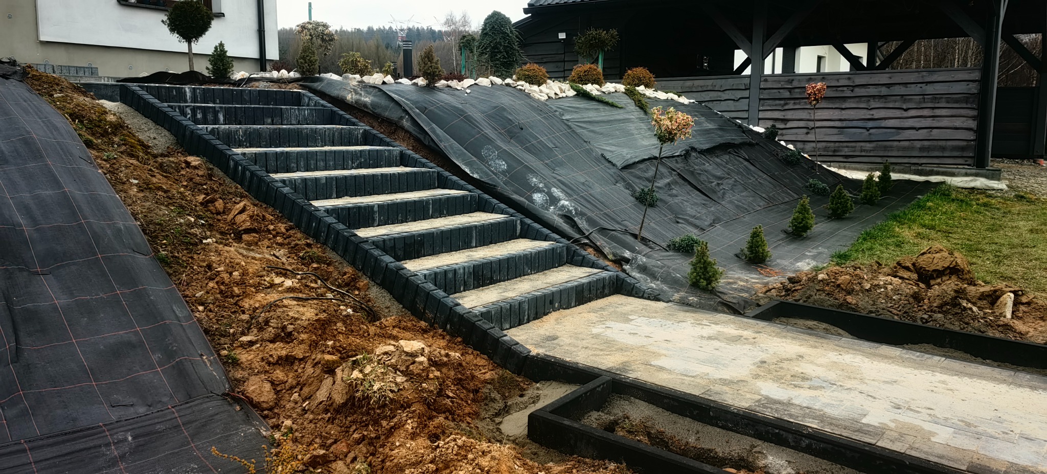 Schody z kostki brukowej prowadzące na wzgórze z agrowłókniną i nasadzeniami, widok z dołu, w tle drewniana altana. Etap prac wykończeniowych.