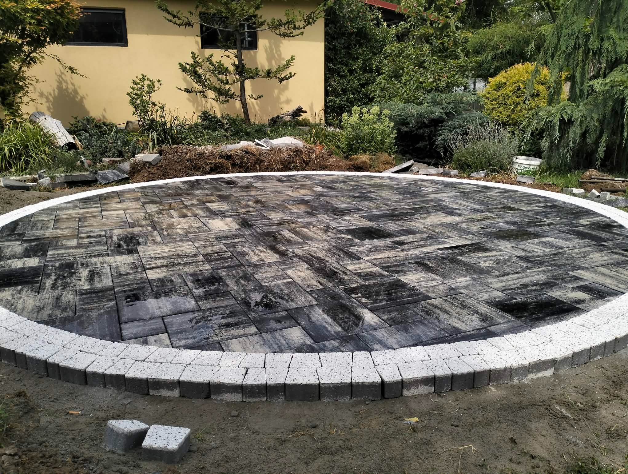 Okrągły plac z ułożonej, szarej kostki brukowej z białym obramowaniem, w tle budynek i zieleń. Widoczne niedokończone prace i przygotowany teren.