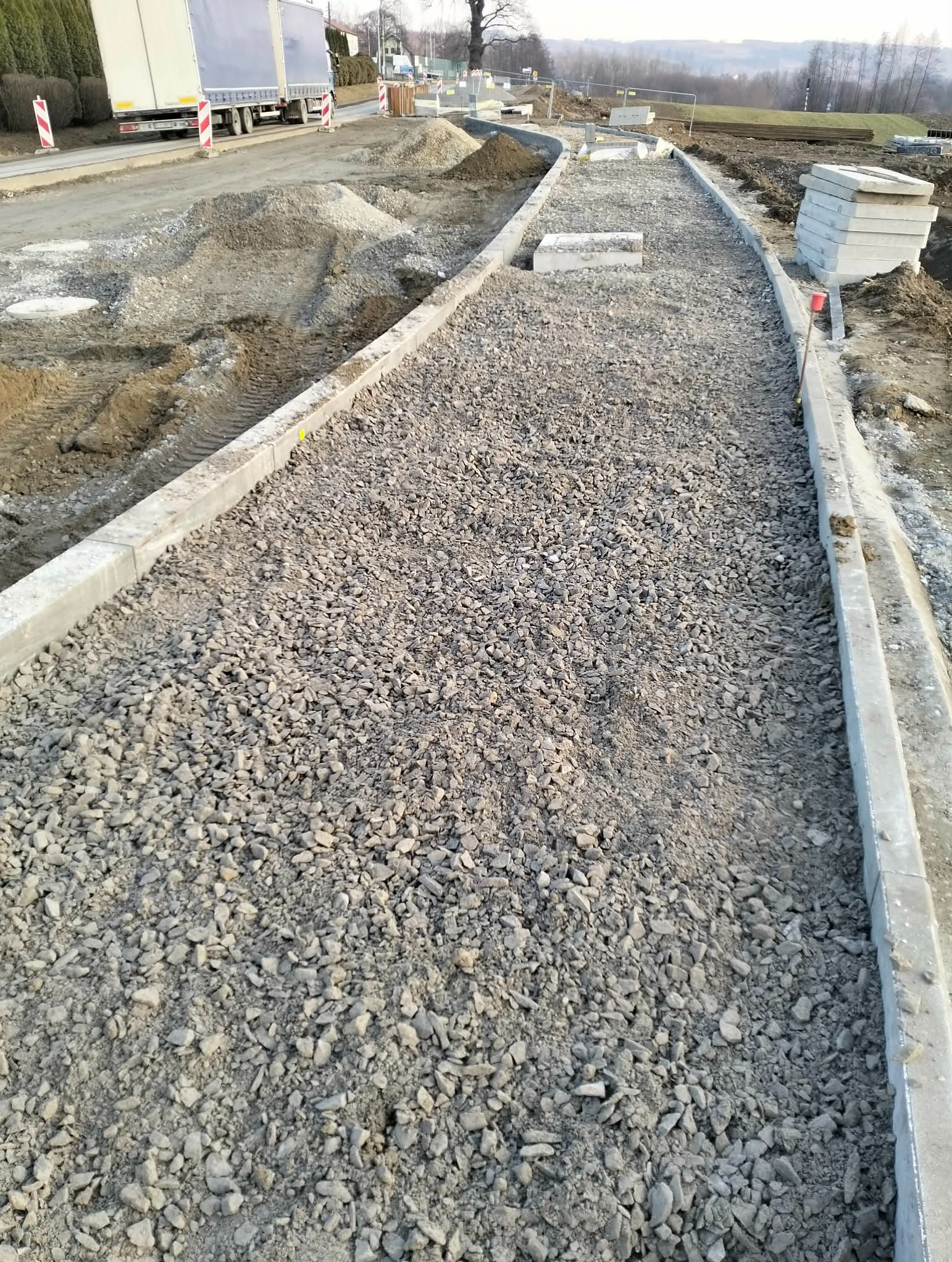 Przygotowany podkład z kruszywa ograniczonego obrzeżami, widoczne hałdy ziemi i ciężarówka w tle, sugerujące prace drogowe lub przygotowanie pod nawierzchnię.