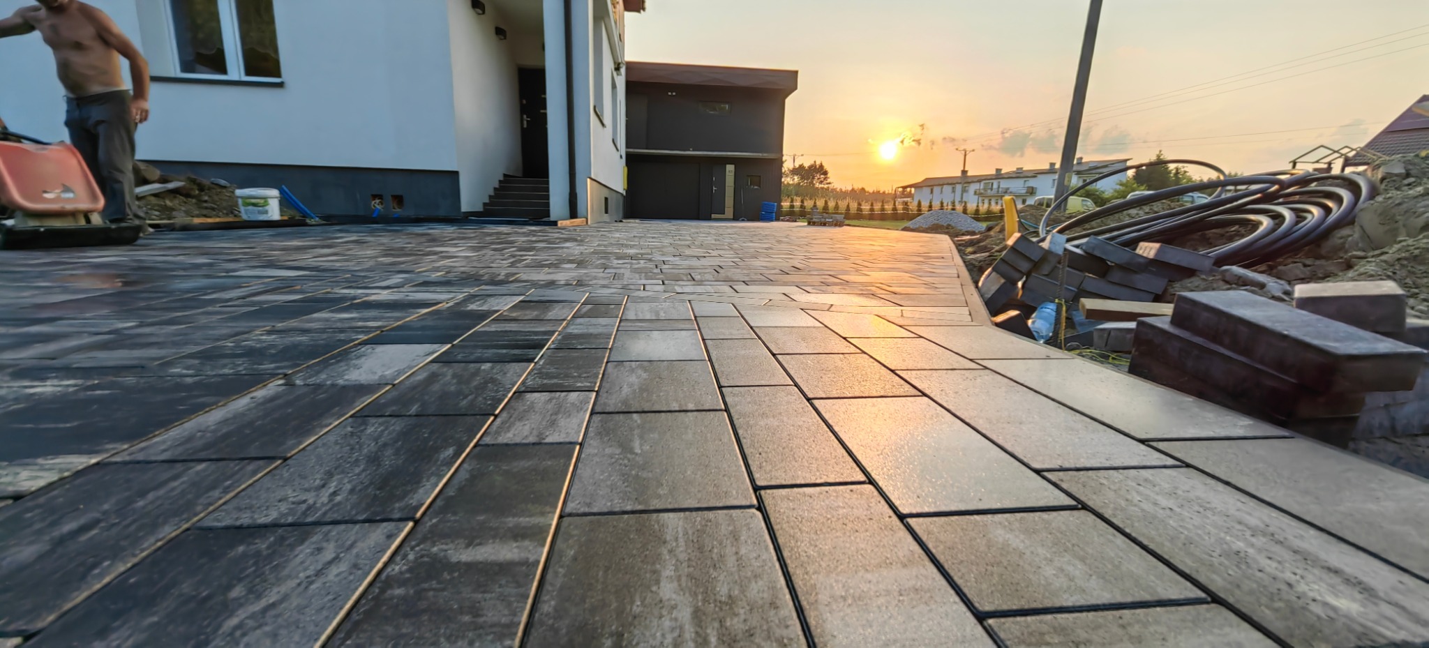 Ułożona, prostokątna nawierzchnia z szarej kostki, z widocznym fragmentem domu, sprzętem budowlanym i zachodzącym słońcem w tle. Kąt ujęcia z dołu podkreśla perspektywę.