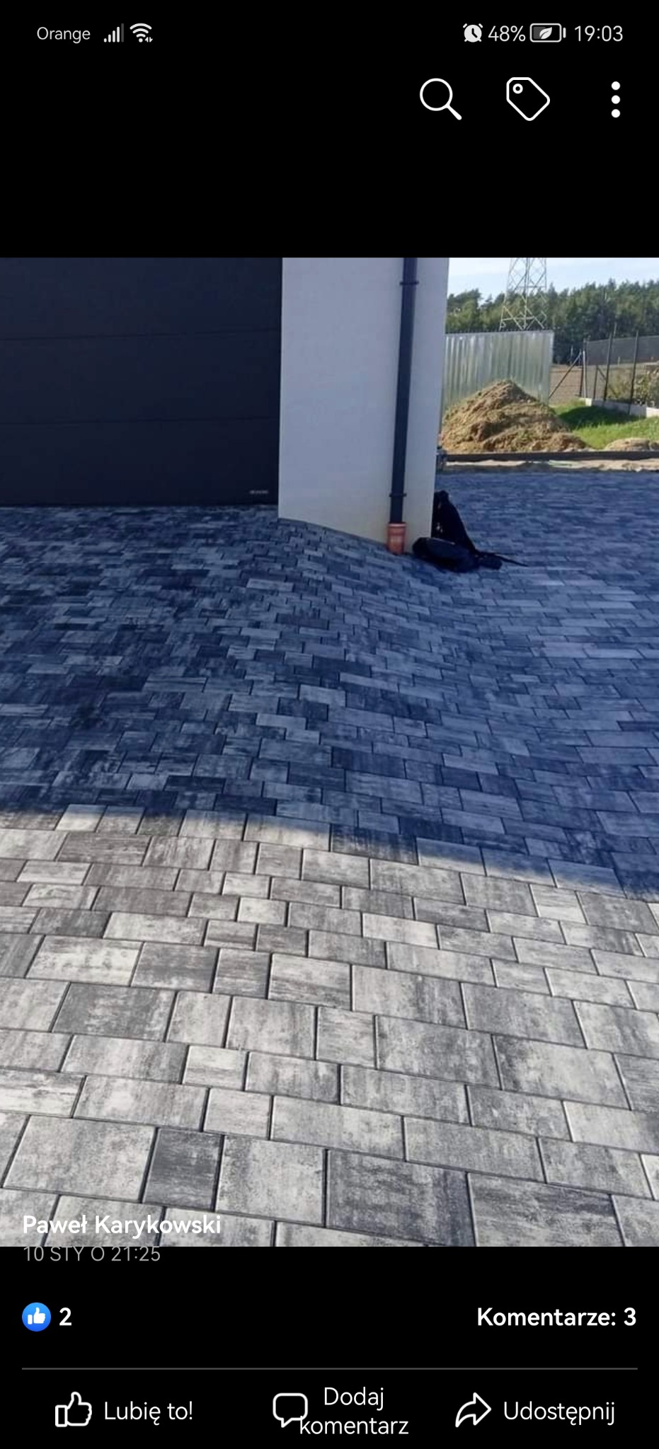 Ułożona, szara kostka brukowa przed budynkiem z ciemną bramą garażową. Widoczny fragment rynny i sterta ziemi w tle. Ujęcie z niskiej perspektywy.