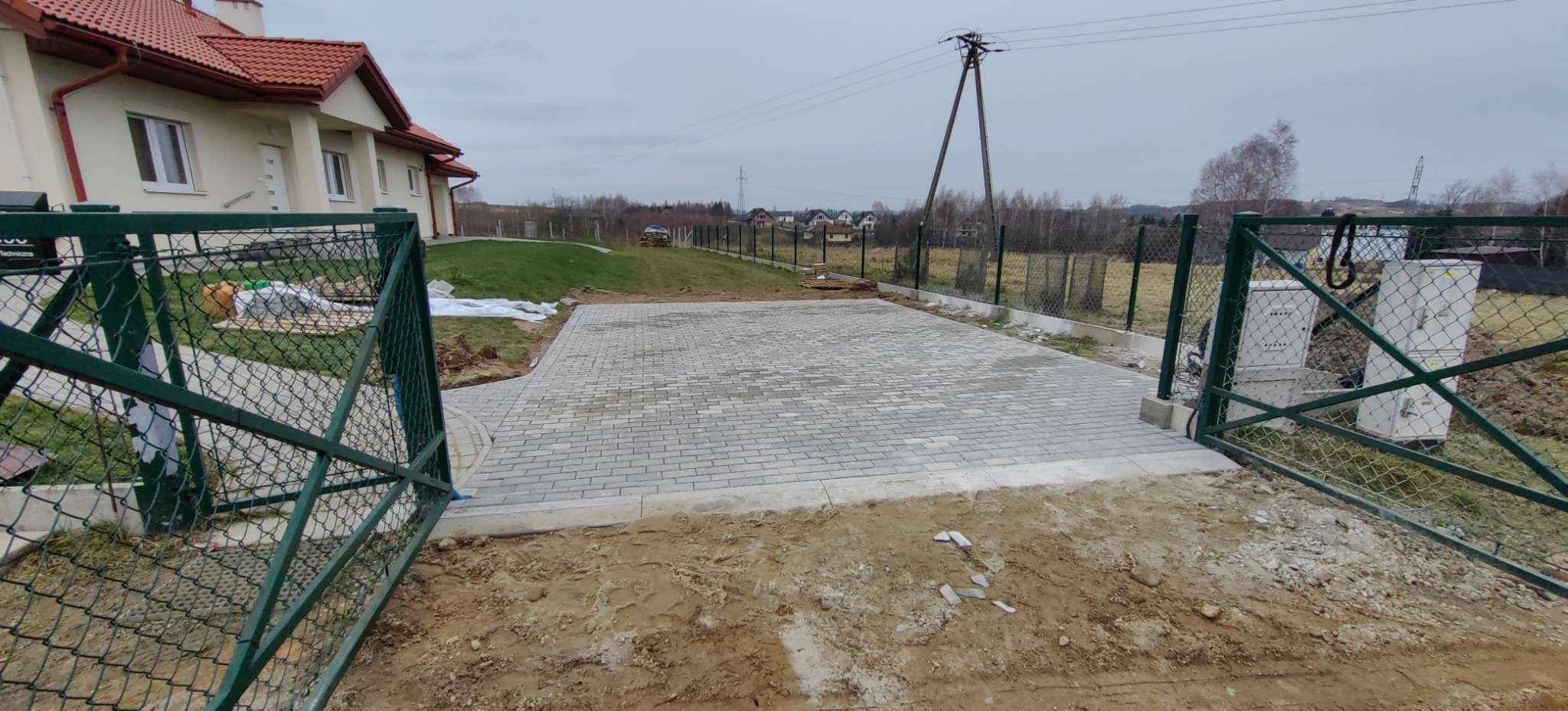 Szara kostka ułożona na podjeździe, widok przez otwartą, zieloną bramę. W tle dom z czerwoną dachówką i zielony trawnik. Słup energetyczny w oddali.