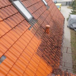 MALVERTO SP. Z O. O. - Częściowo wyczyszczony, pomarańczowy dach z dachówki ceramicznej, widoczne dwa okna dachowe i komin. Połowa dachu czysta, druga pokryta mchem i zabrudzeniami.