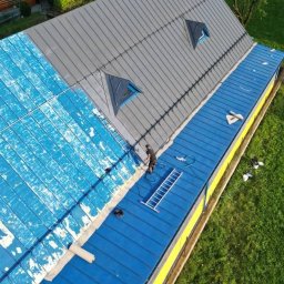 MALVERTO SP. Z O. O. - Prace na dachu: część dachu szara, część niebieska, pracownik na drabinie. Widok z góry ukazuje postęp prac na tle zieleni.