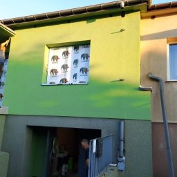 MALVERTO SP. Z O. O. - Malowanie elewacji domu na zielono. Pracownik na drabinie maluje fragment pod dachem. Okno zabezpieczone folią. Widoczny fragment elewacji przed malowaniem.