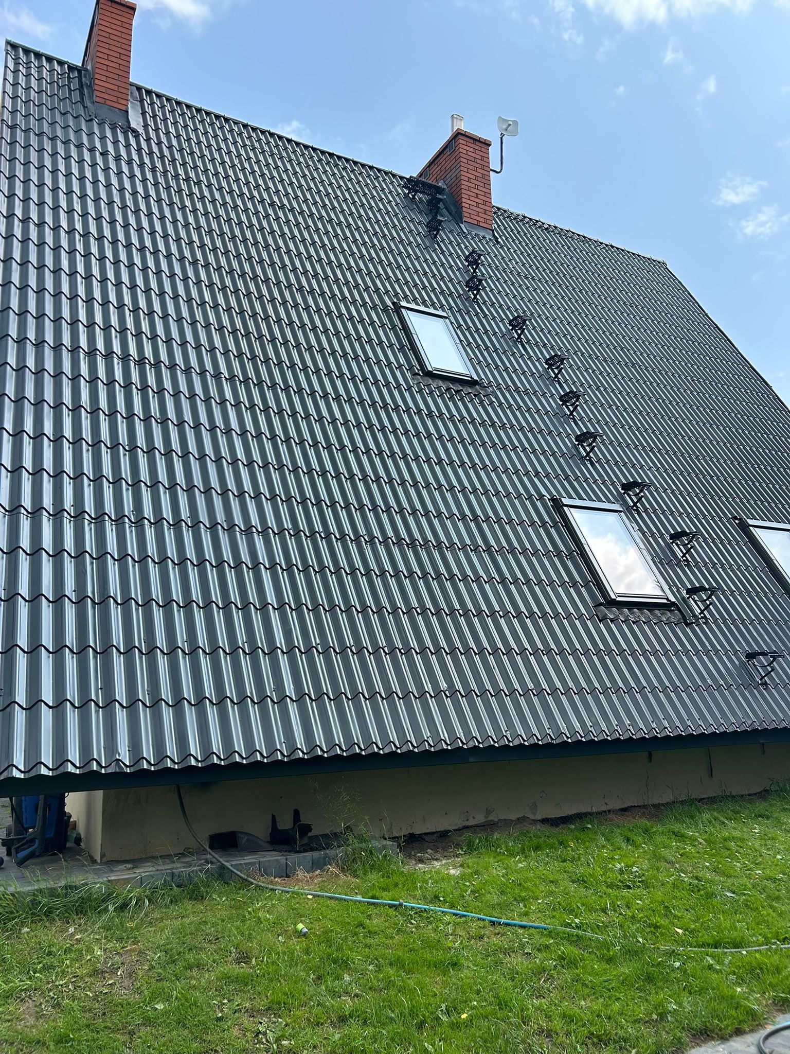 Ciemny, metalowy dach z oknami połaciowymi i ceglanymi kominami na tle błękitnego nieba. Widoczne drabinki dachowe i fragment trawnika.