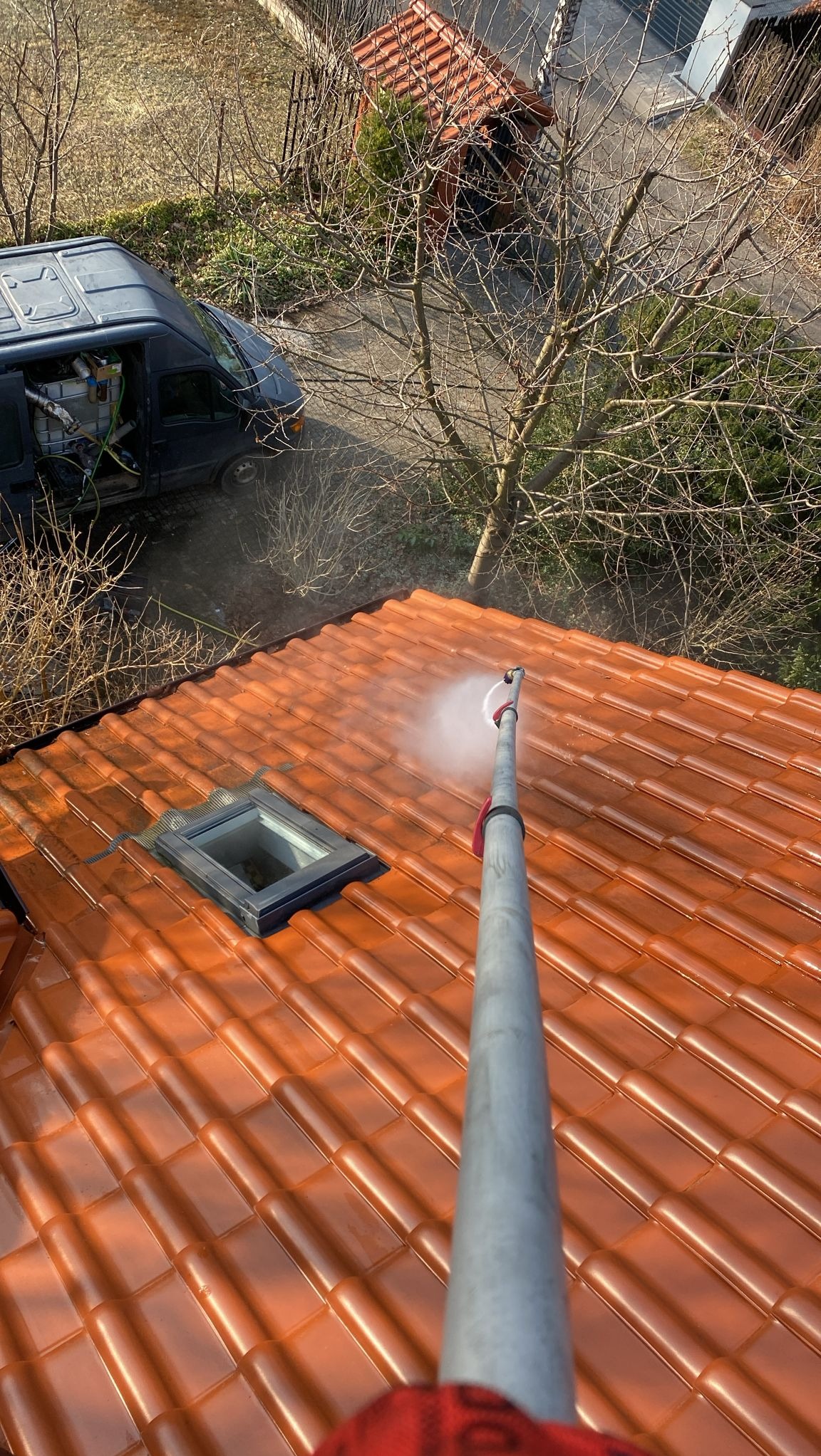 Pomarańczowy dach z dachówki ceramicznej czyszczony lancą ciśnieniową. Widoczny fragment okna dachowego, samochód dostawczy i drzewa w tle. Ujęcie z góry.