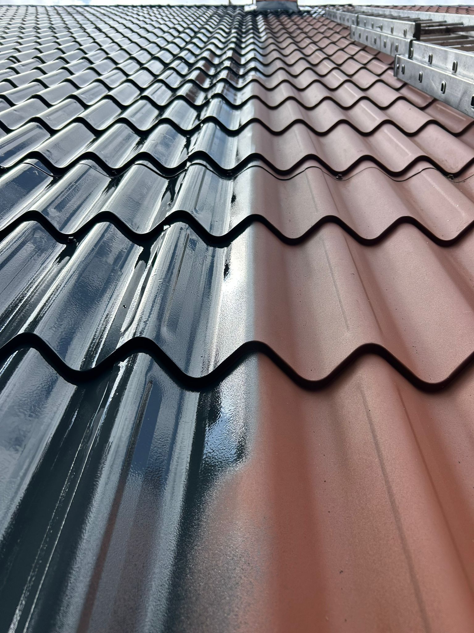 Częściowo pomalowany dach z blachodachówki w zbliżeniu, z widoczną drabiną. Połowa dachu w kolorze grafitowym, druga połowa w kolorze ceglastym. Ukazany efekt przed i po malowaniu.