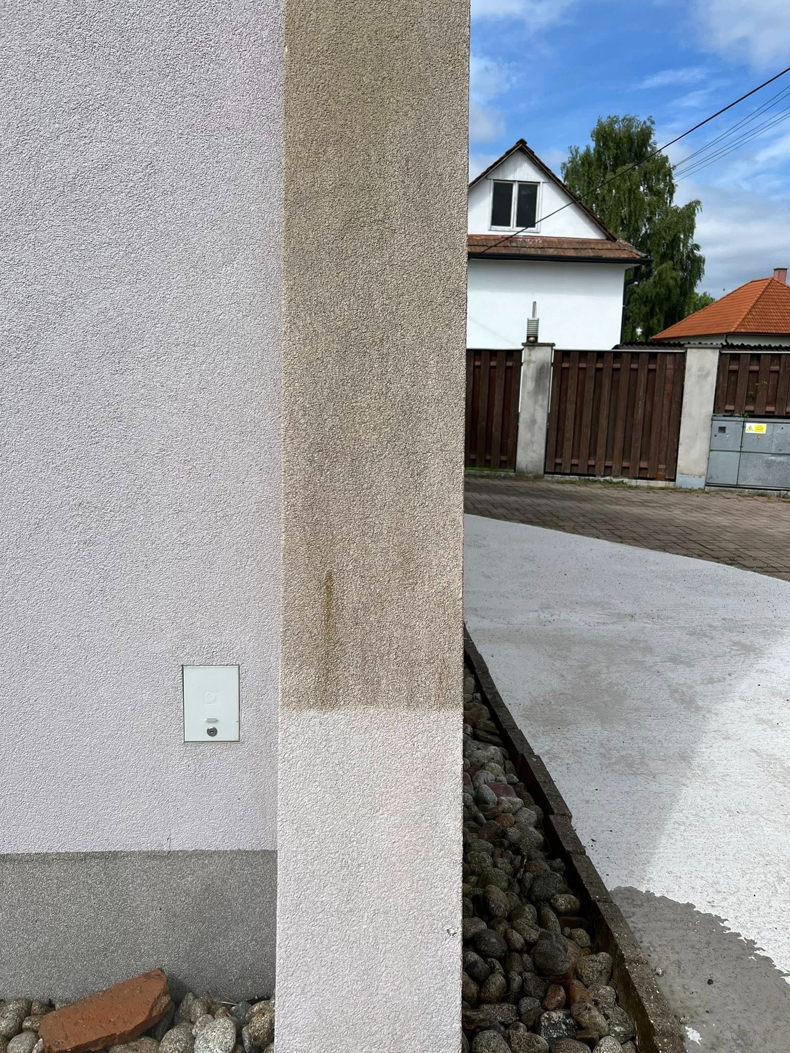 Fragment elewacji budynku z widocznym efektem czyszczenia: część ściany umyta, część zabrudzona. W tle dom i brama wjazdowa.