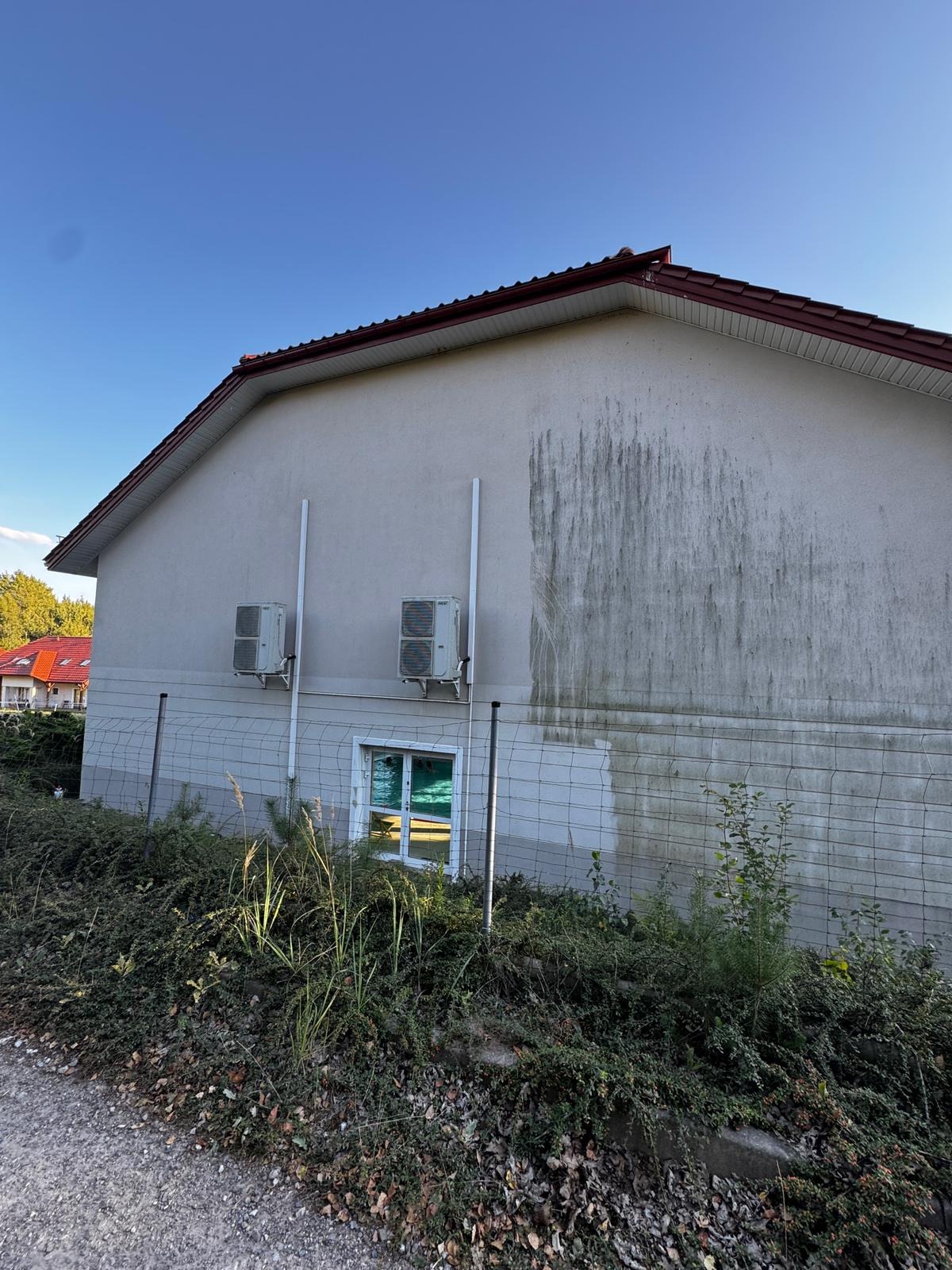 Ściana budynku z widocznymi zabrudzeniami i śladami po myciu, dwa klimatyzatory na elewacji, okno z zielonkawą szybą, siatka ogrodzeniowa i zarośla.