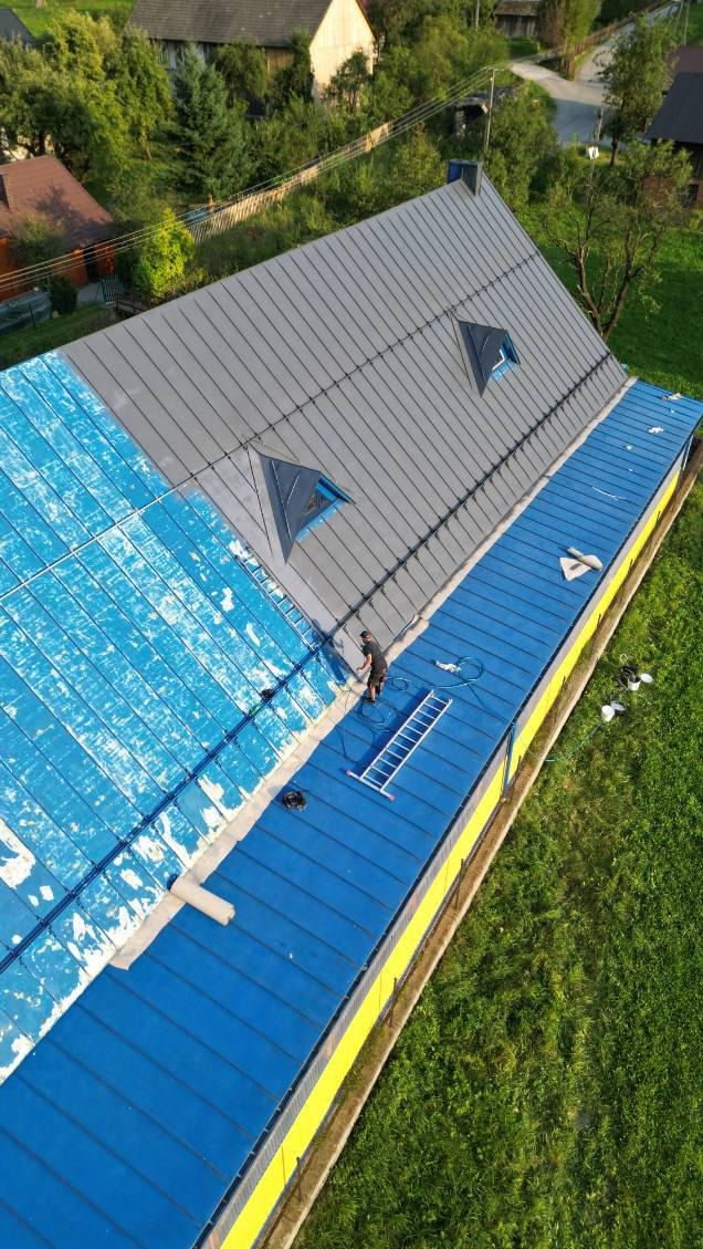 Prace na dachu: część dachu szara, część niebieska, pracownik na drabinie. Widok z góry ukazuje postęp prac na tle zieleni.