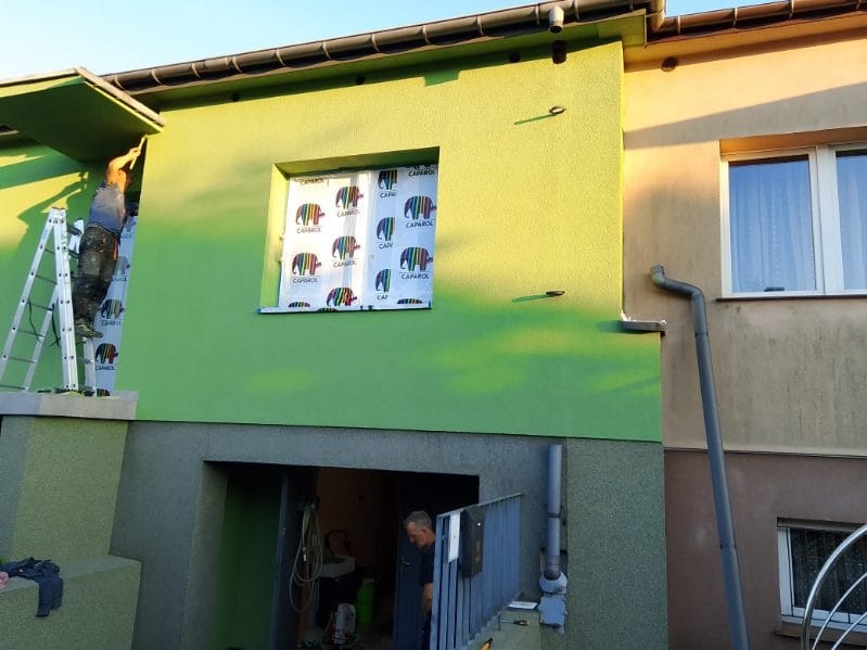 Malowanie elewacji domu na zielono. Pracownik na drabinie maluje fragment pod dachem. Okno zabezpieczone folią. Widoczny fragment elewacji przed malowaniem.