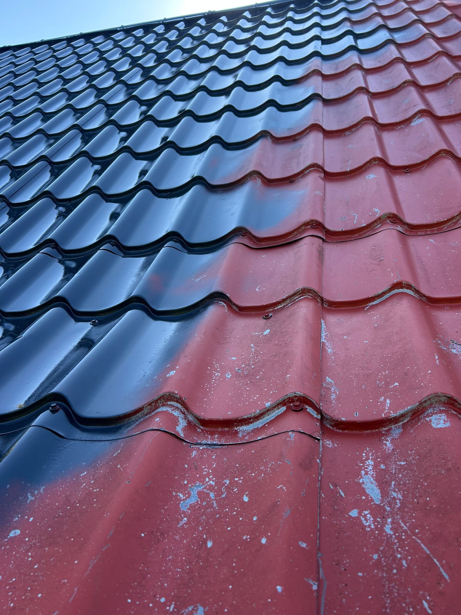 Częściowo pomalowany dach z blachodachówki w kolorze czerwonym i czarnym. Widoczne ślady starej farby. Ujęcie z dołu, ukazujące strukturę i detale pokrycia.