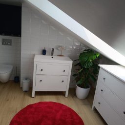 Mateusz Turek - Jasna łazienka na poddaszu ze skośnym sufitem, białe meble z czarnymi gałkami, czerwony dywanik i roślina w doniczce. Białe płytki na ścianach.