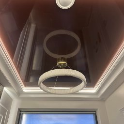 Mateusz Turek - Błyszczący, bordowy sufit napinany z okrągłą lampą LED i podświetleniem obwodowym. Okno zabezpieczone folią malarską.