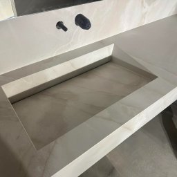 Mateusz Turek - Wykończona, beżowa umywalka wpuszczana w blat z kamienia. Widoczne czarne elementy armatury. Ujęcie z góry, z fragmentem buta w prawym dolnym rogu.