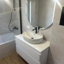 Mateusz Turek - Nowoczesna łazienka po remoncie: owalne lustro, biała umywalka nablatowa z czarną armaturą, szare płytki i drewnopodobna podłoga. Minimalistyczny design.