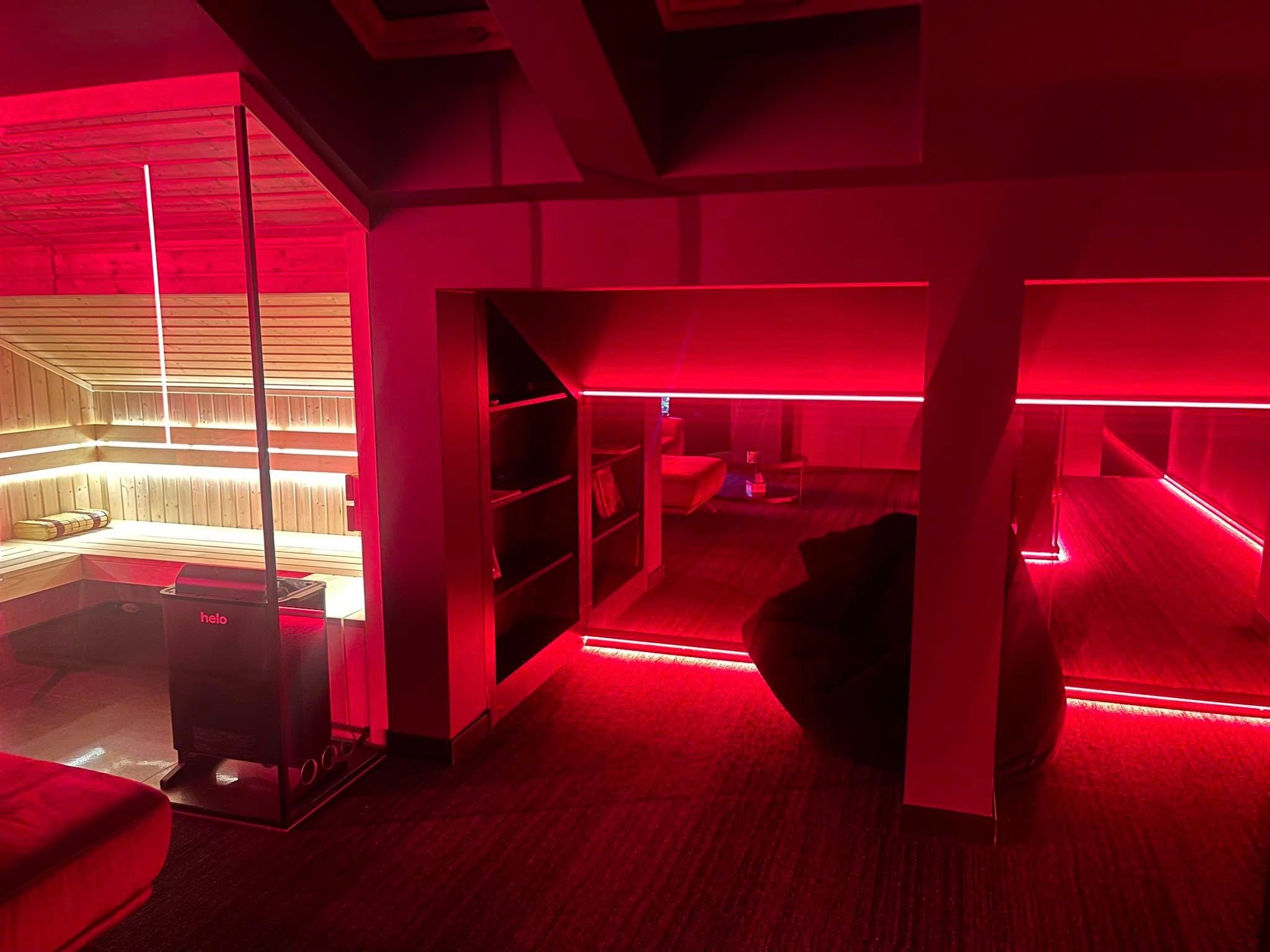 Nowoczesna sauna z piecem 'helo' i oświetleniem LED w intensywnym czerwonym kolorze, widok na strefę relaksu z pufem i regałem.