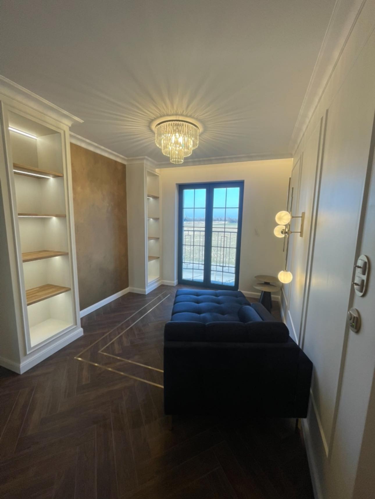 Eleganckie wykończenie wnętrza w Warszawie: parkiet w jodełkę z metalowymi listwami, zabudowane regały z oświetleniem i dekoracyjna ściana w odcieniach brązu.