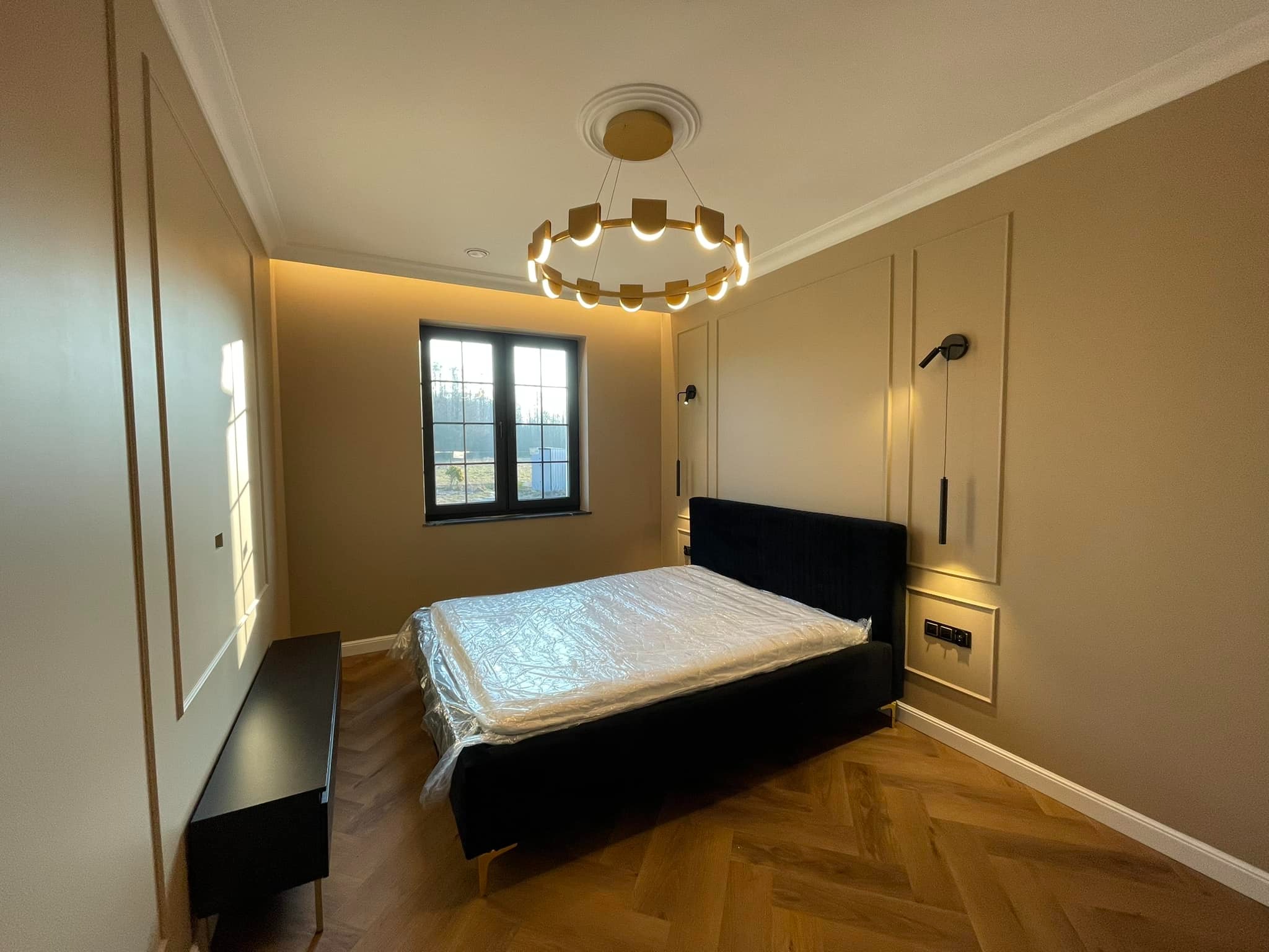 Elegancka sypialnia z czarnym łóżkiem i parkietem w jodełkę. Ściany w odcieniach beżu z dekoracyjnymi listwami. Okrągła lampa sufitowa i minimalistyczne kinkiety.