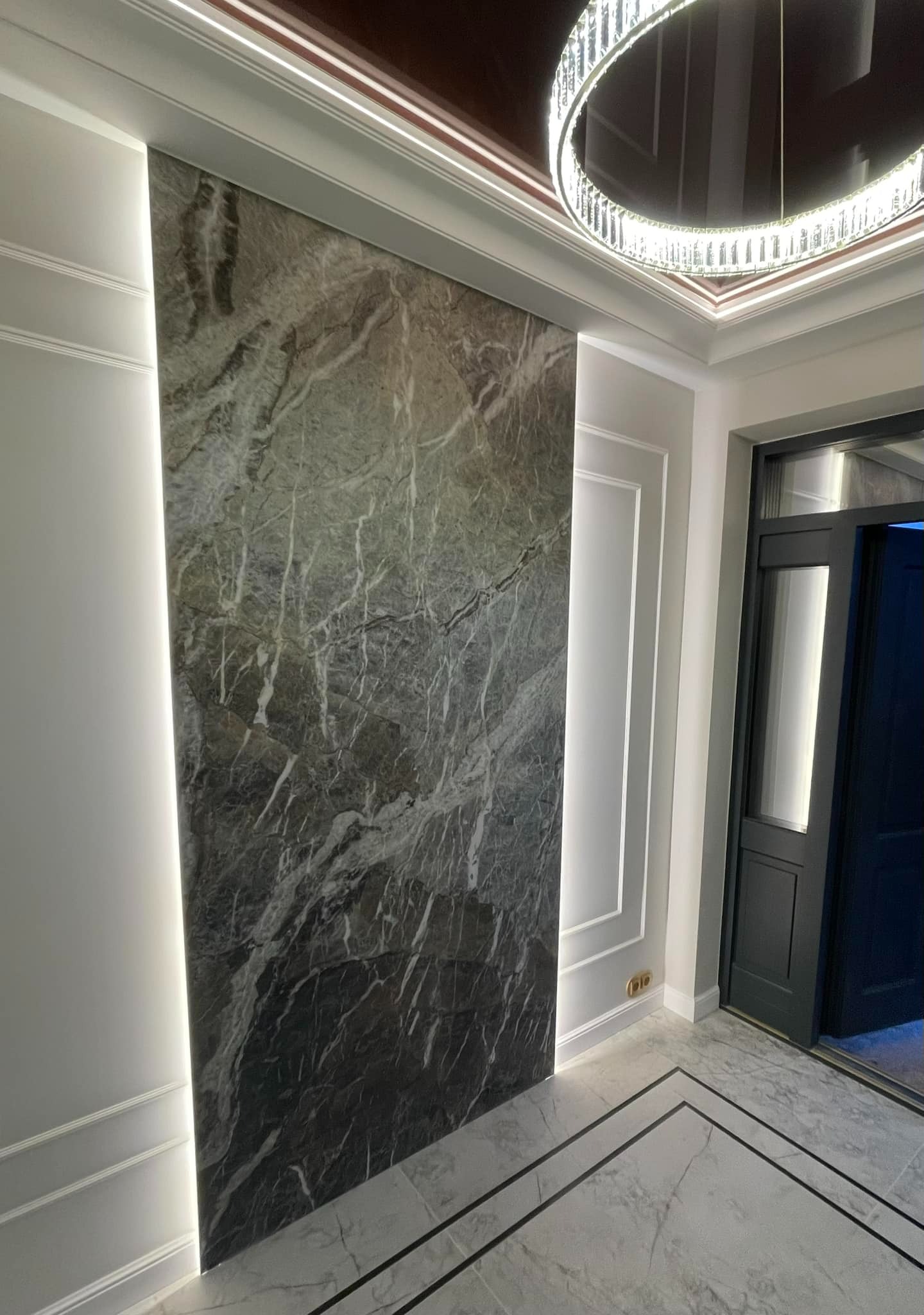 Eleganckie wykończenie wnętrza: marmurowa ściana z podświetleniem LED, dekoracyjne listwy ścienne, marmurowa podłoga z czarnymi akcentami i nowoczesny żyrandol.