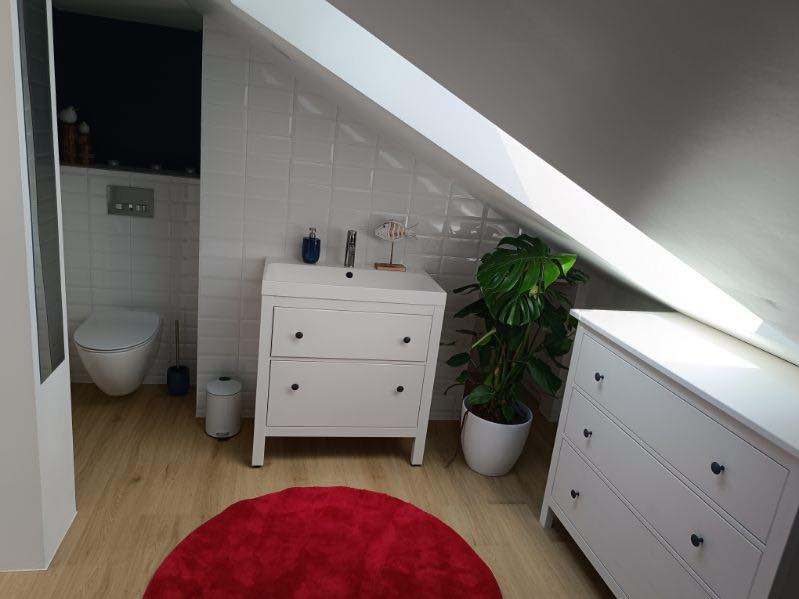 Jasna łazienka na poddaszu ze skośnym sufitem, białe meble z czarnymi gałkami, czerwony dywanik i roślina w doniczce. Białe płytki na ścianach.