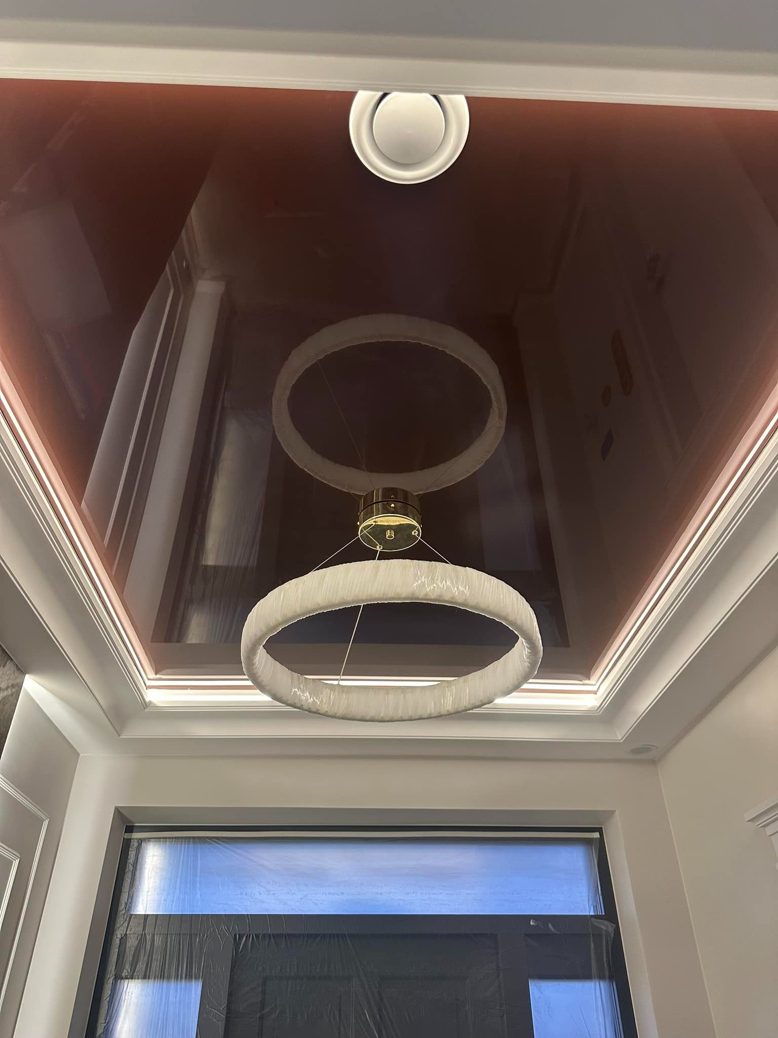 Błyszczący, bordowy sufit napinany z okrągłą lampą LED i podświetleniem obwodowym. Okno zabezpieczone folią malarską.