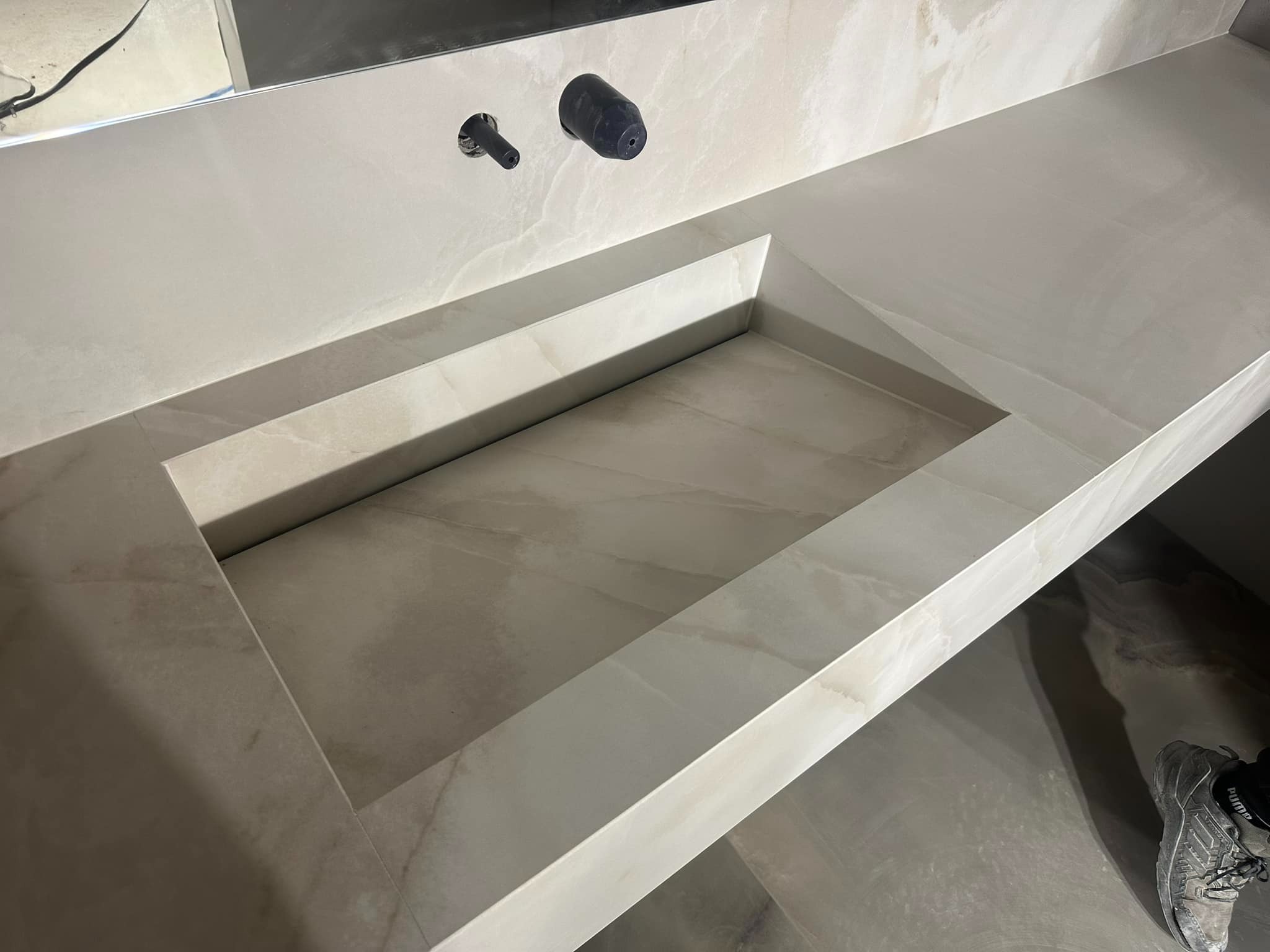 Wykończona, beżowa umywalka wpuszczana w blat z kamienia. Widoczne czarne elementy armatury. Ujęcie z góry, z fragmentem buta w prawym dolnym rogu.