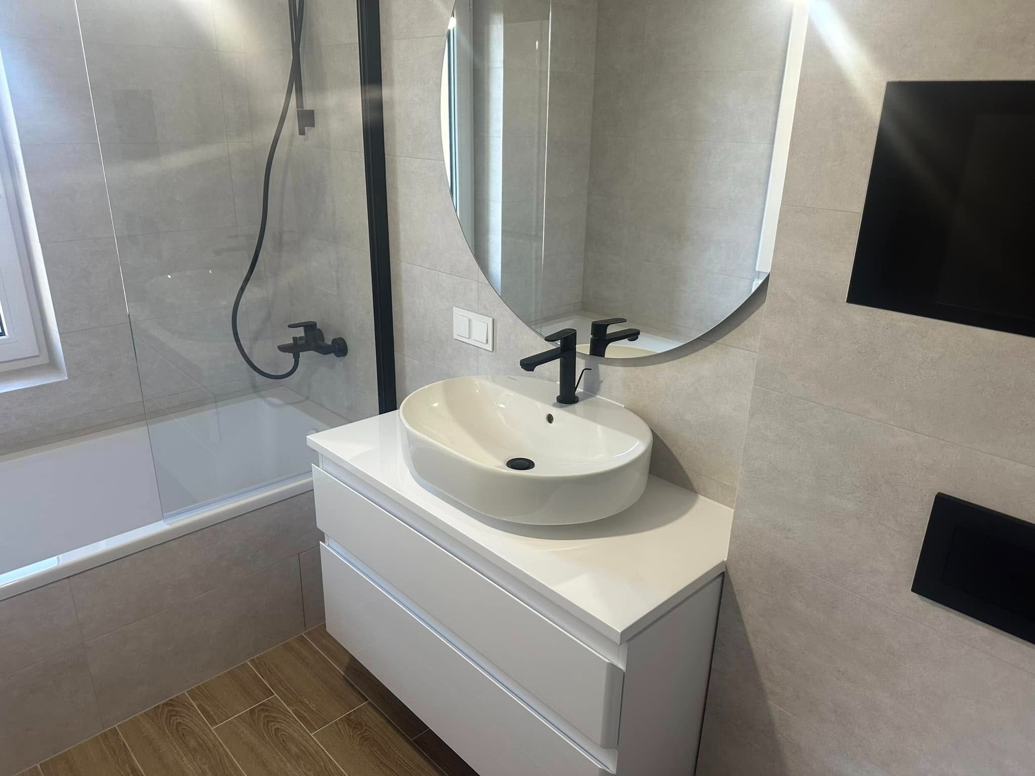 Nowoczesna łazienka po remoncie: owalne lustro, biała umywalka nablatowa z czarną armaturą, szare płytki i drewnopodobna podłoga. Minimalistyczny design.