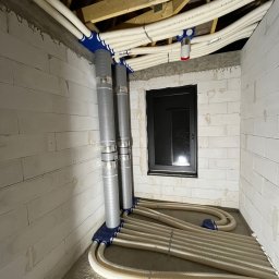 RESMONT - Instalacja wentylacyjna z rurami w surowym wnętrzu budynku z oknem. Widoczne elementy montażowe i podłączenia. Szare ściany z betonu komórkowego.