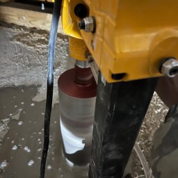 RESMONT - Żółta wiertnica rdzeniowa w trakcie pracy, wiercąca otwór w betonowej posadzce. Wokół wiertła woda, widoczne elementy konstrukcyjne i przewody.