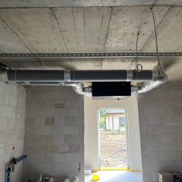 RESMONT - Wnętrze surowego domu z widoczną instalacją rekuperacji pod betonowym stropem. Szare rury z srebrną taśmą, czarna skrzynka rekuperatora. W tle widok na zewnątrz przez otwór okienny.