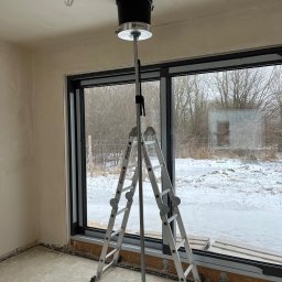 RESMONT - Wnętrze w stanie deweloperskim z drabiną i narzędziem do montażu wentylacji sufitowej. Widoczne okno z zimowym krajobrazem. Przewody elektryczne wystają z sufitu.