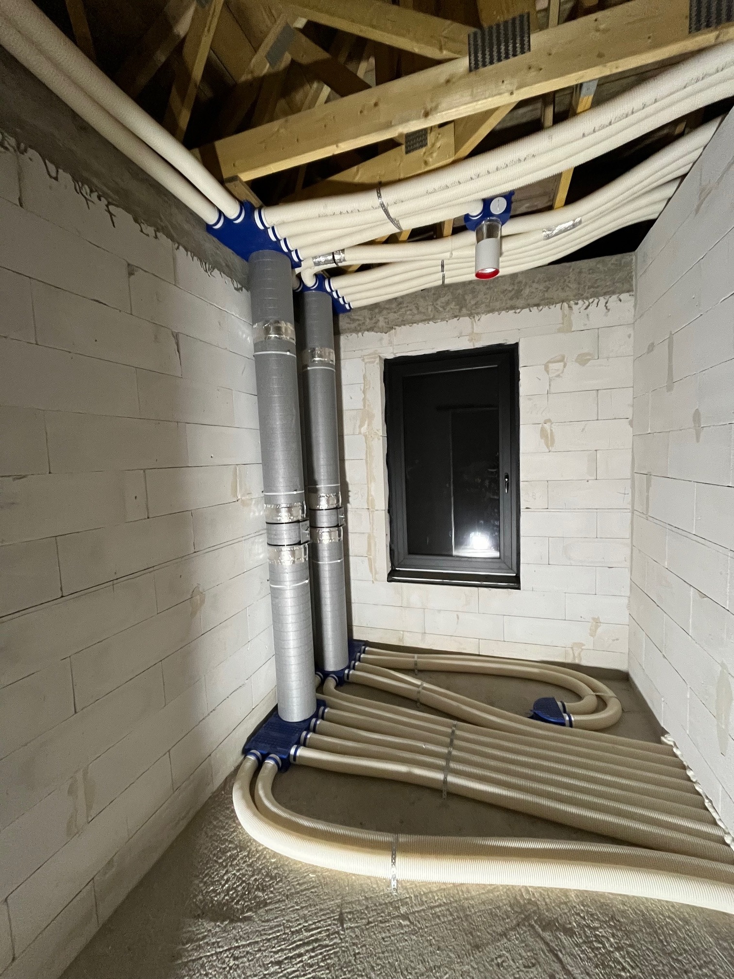 Instalacja wentylacyjna z rurami w surowym wnętrzu budynku z oknem. Widoczne elementy montażowe i podłączenia. Szare ściany z betonu komórkowego.