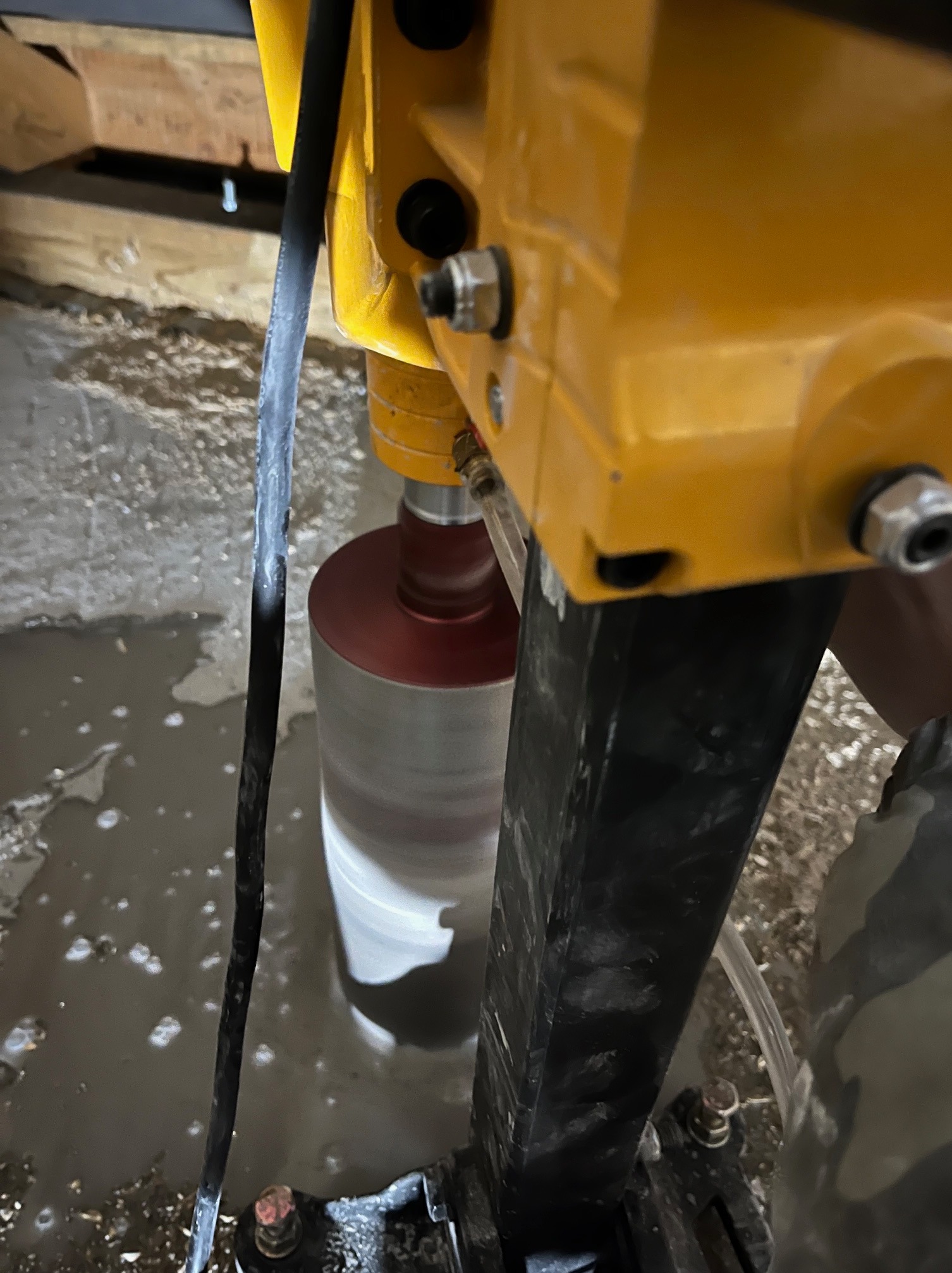 Żółta wiertnica rdzeniowa w trakcie pracy, wiercąca otwór w betonowej posadzce. Wokół wiertła woda, widoczne elementy konstrukcyjne i przewody.