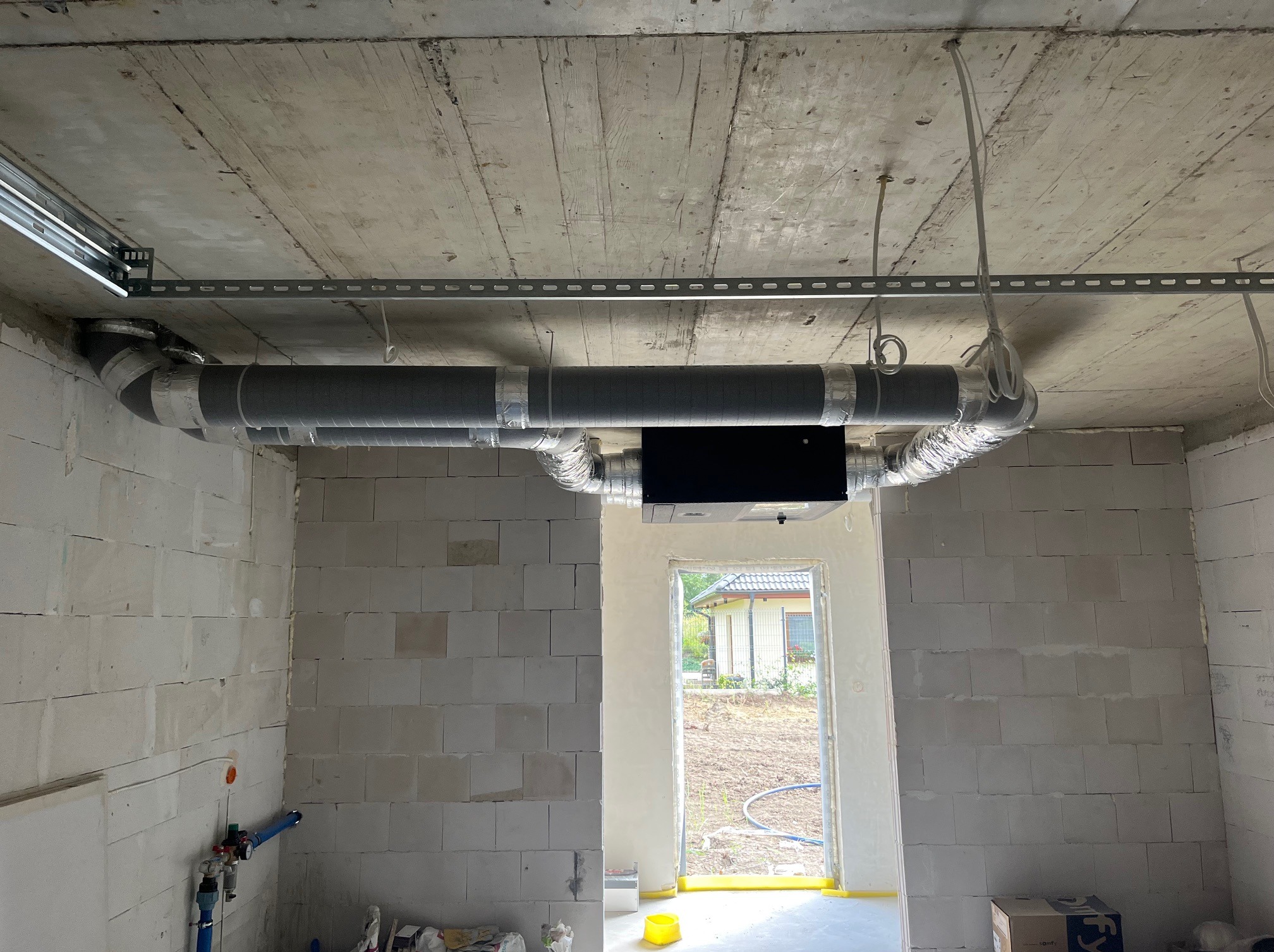 Wnętrze surowego domu z widoczną instalacją rekuperacji pod betonowym stropem. Szare rury z srebrną taśmą, czarna skrzynka rekuperatora. W tle widok na zewnątrz przez otwór okienny.