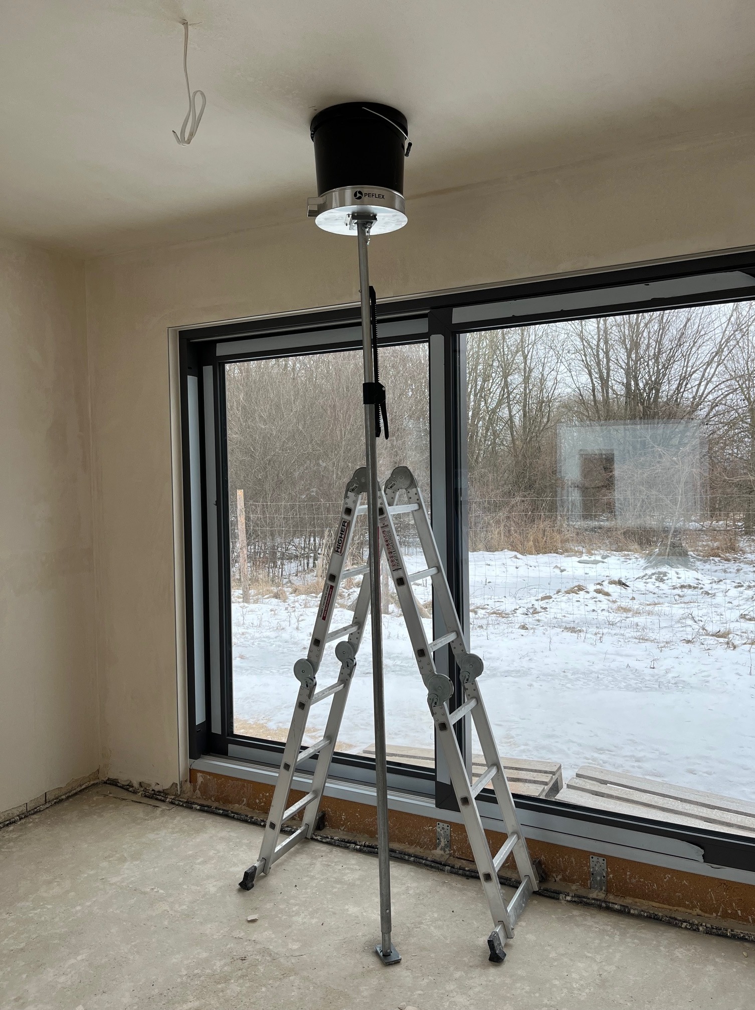 Wnętrze w stanie deweloperskim z drabiną i narzędziem do montażu wentylacji sufitowej. Widoczne okno z zimowym krajobrazem. Przewody elektryczne wystają z sufitu.