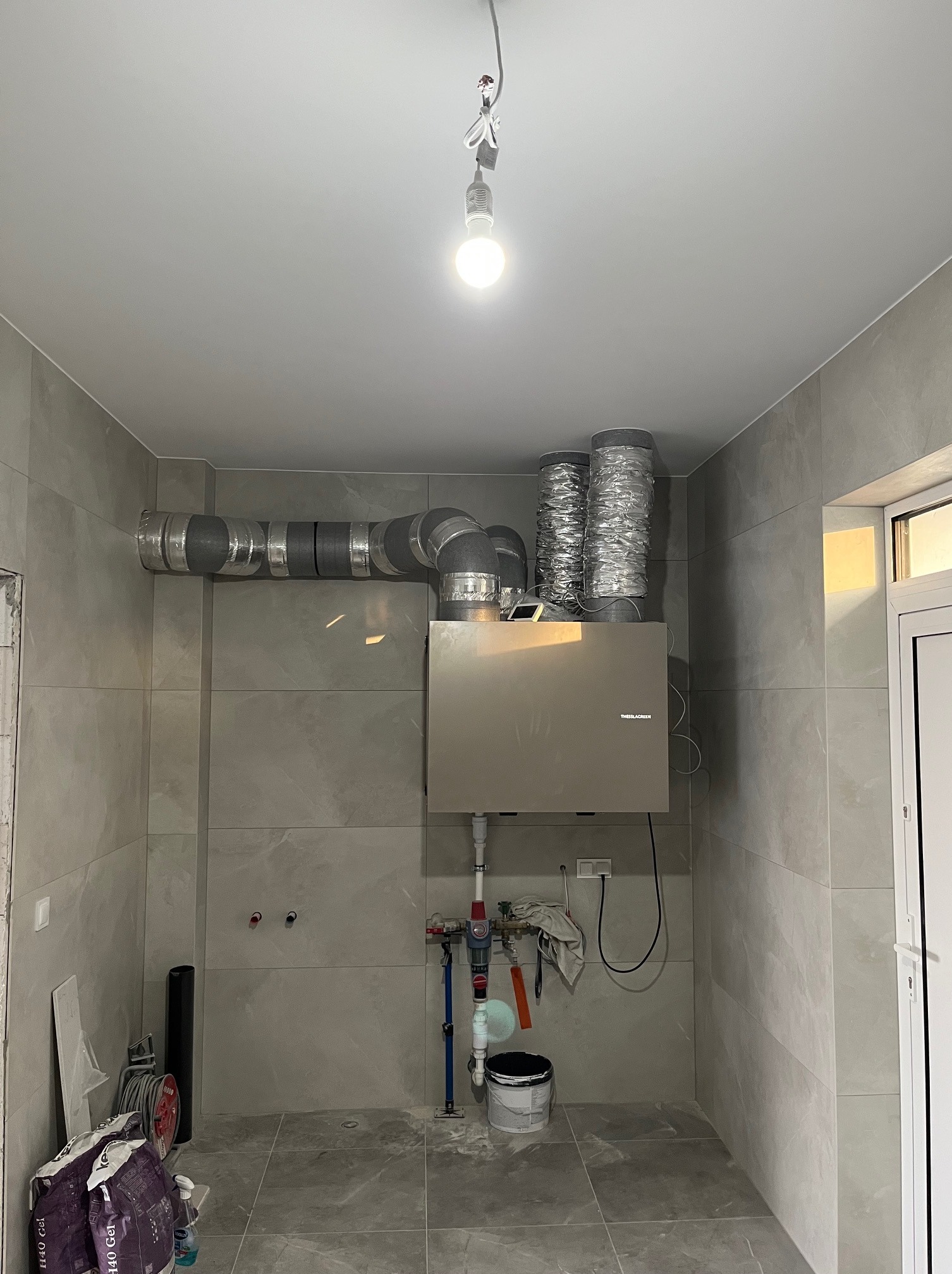 Szara jednostka rekuperacji z podłączonymi przewodami wentylacyjnymi w pomieszczeniu wykończonym płytkami. Widoczne elementy instalacji hydraulicznej i akcesoria malarskie.