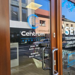 Centrum Serwisowe - Wejście do serwisu Centrum z godzinami otwarcia i ofertą: telefony, tablety i inne urządzenia. Na szybie logo firmy i grafiki w niebieskim kolorze.