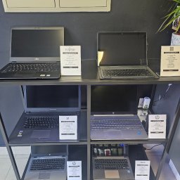 Centrum Serwisowe - Ekspozycja laptopów różnych marek (Fujitsu, Microsoft, Dream Machines, HP, Asus) na metalowym regale w sklepie. Widoczne specyfikacje i ceny.