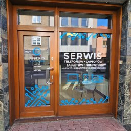 Centrum Serwisowe - Wejście do serwisu elektroniki z naklejkami informacyjnymi na szybie: telefony, laptopy, tablety, komputery. Widoczne logo firmy i godziny otwarcia.