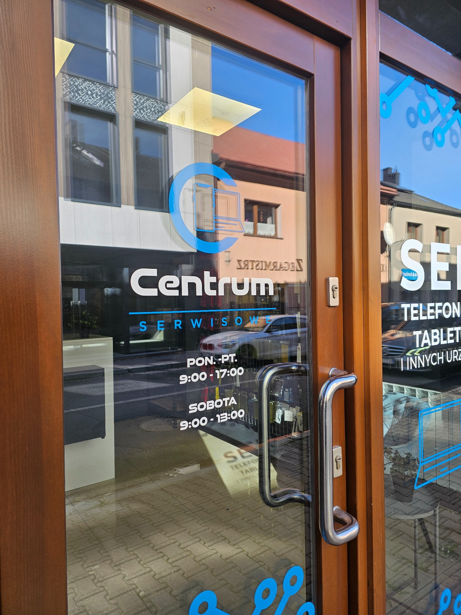 Wejście do serwisu Centrum z godzinami otwarcia i ofertą: telefony, tablety i inne urządzenia. Na szybie logo firmy i grafiki w niebieskim kolorze.