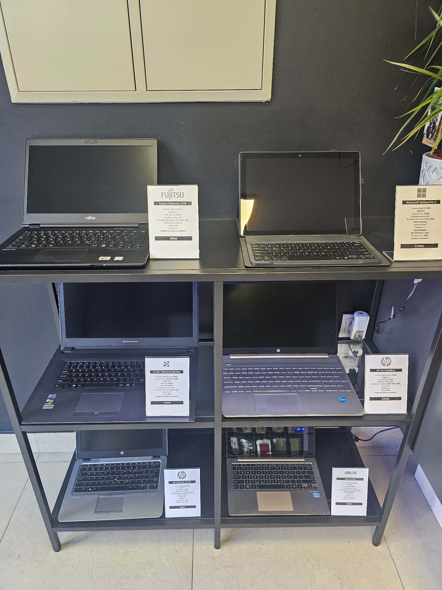 Ekspozycja laptopów różnych marek (Fujitsu, Microsoft, Dream Machines, HP, Asus) na metalowym regale w sklepie. Widoczne specyfikacje i ceny.
