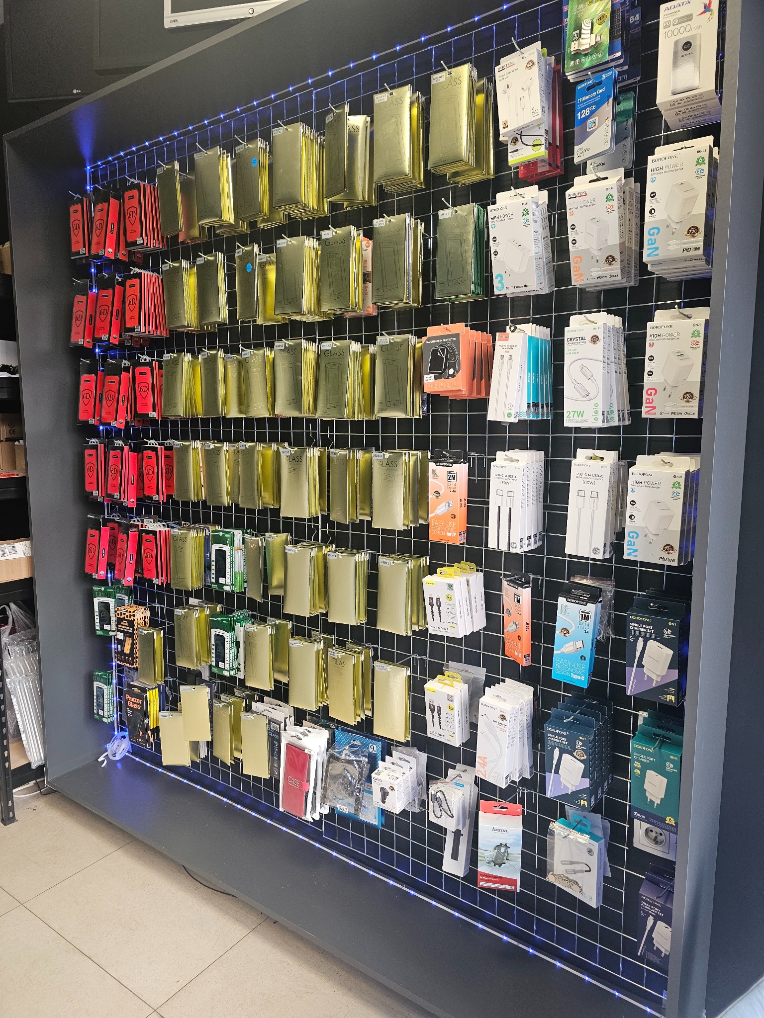 Ekspozycja akcesoriów GSM: szkła hartowane, ładowarki, kable USB i słuchawki. Różnorodność produktów na metalowej siatce z podświetleniem LED.