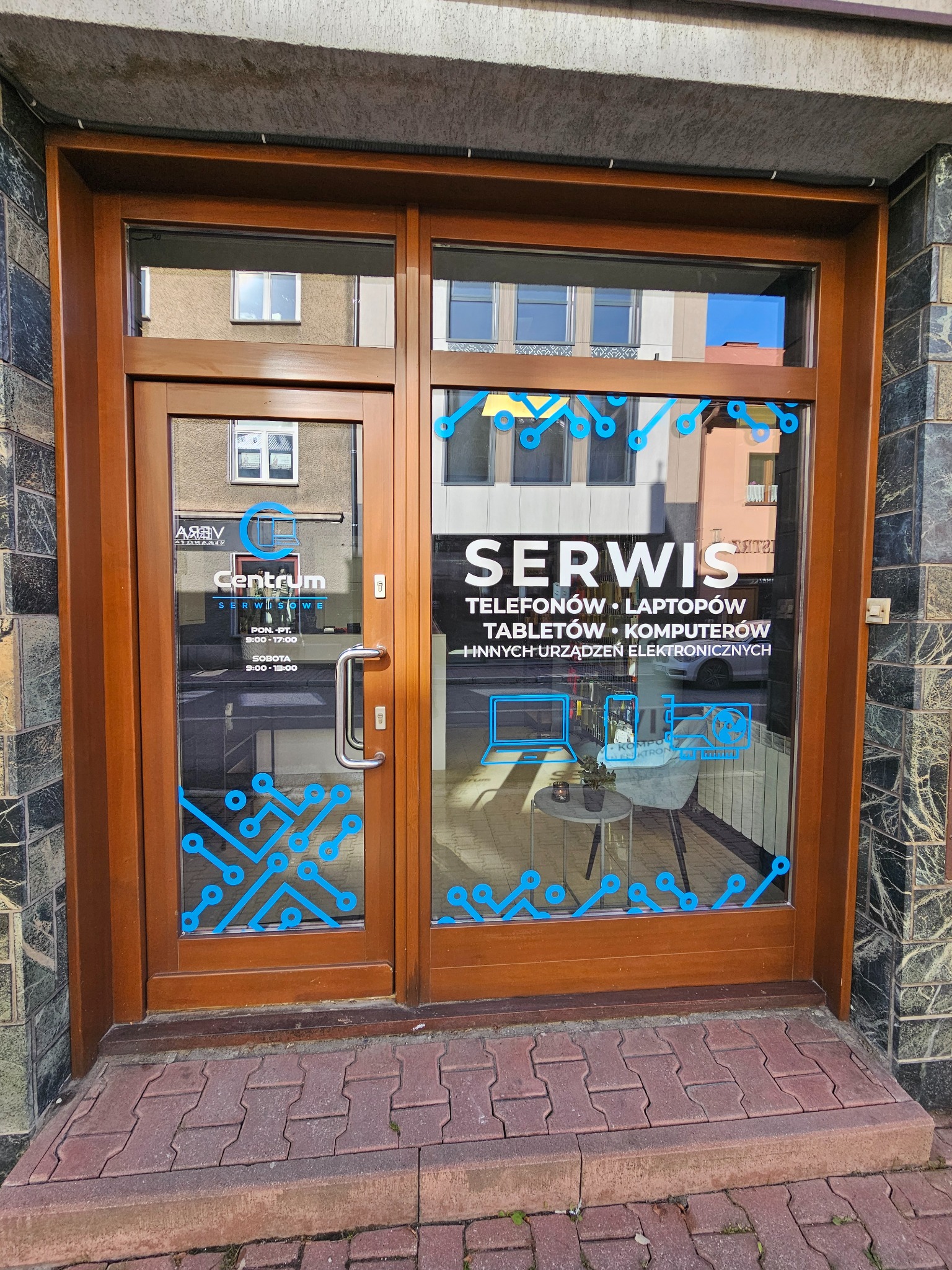 Wejście do serwisu elektroniki z naklejkami informacyjnymi na szybie: telefony, laptopy, tablety, komputery. Widoczne logo firmy i godziny otwarcia.