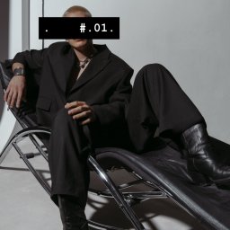 Dominik Kowalczyk - Osoba w czarnym garniturze i kowbojkach siedzi na chromowanym krześle. Twarz zasłonięta czarnym paskiem z napisem '.#.01.'. Srebrna biżuteria.