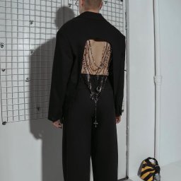 Dominik Kowalczyk - Osoba w czarnym, awangardowym garniturze z wycięciem na plecach, ozdobionym łańcuchami i krzyżami. Widok z tyłu, minimalistyczne tło, torba w paski leży na podłodze.