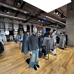 Dominik Kowalczyk - Jasne wnętrze sklepu odzieżowego z manekinami prezentującymi najnowsze kolekcje. Widoczne ubrania na wieszakach i drewniana podłoga. Oświetlenie sufitowe.