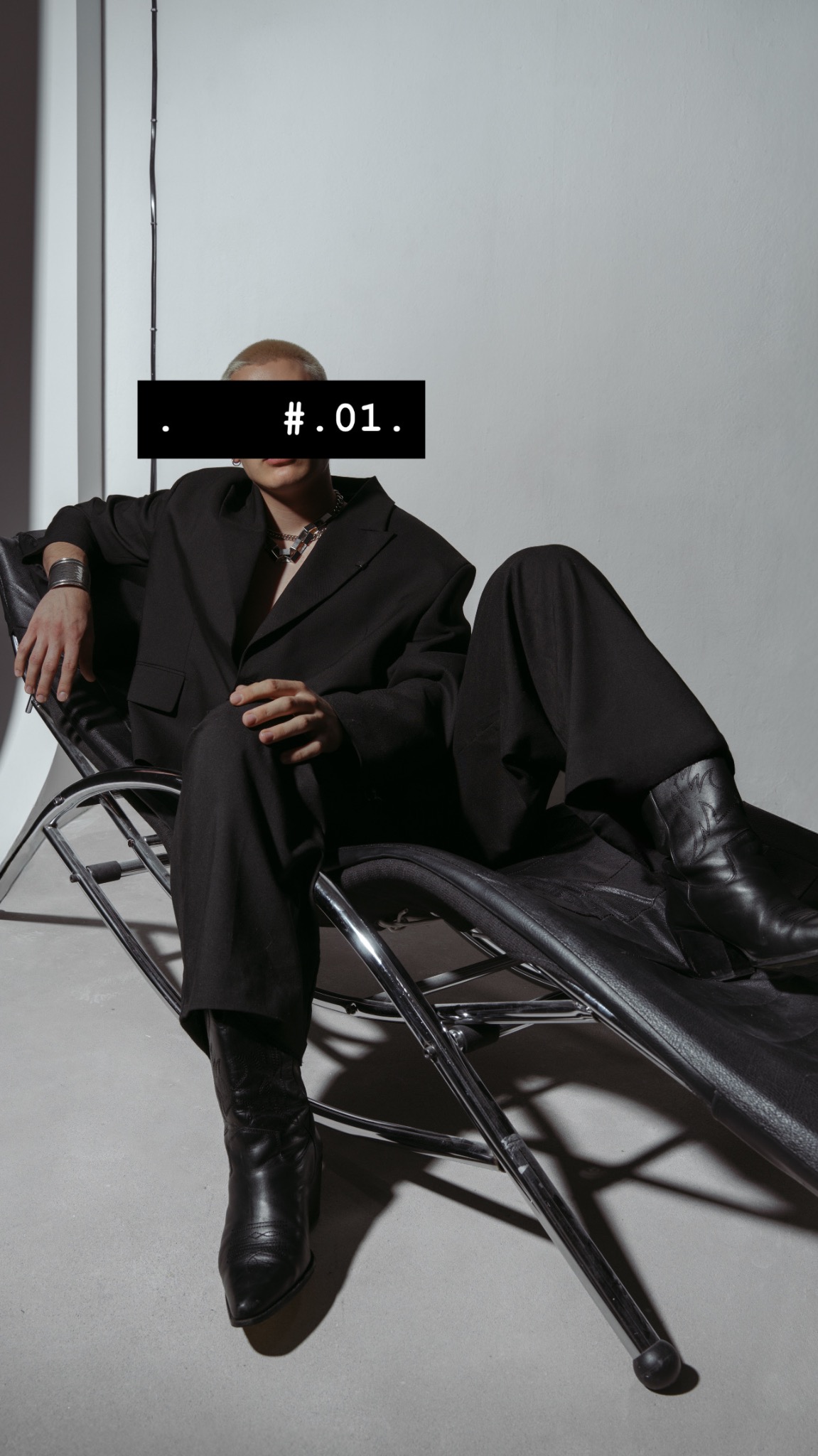 Osoba w czarnym garniturze i kowbojkach siedzi na chromowanym krześle. Twarz zasłonięta czarnym paskiem z napisem '.#.01.'. Srebrna biżuteria.