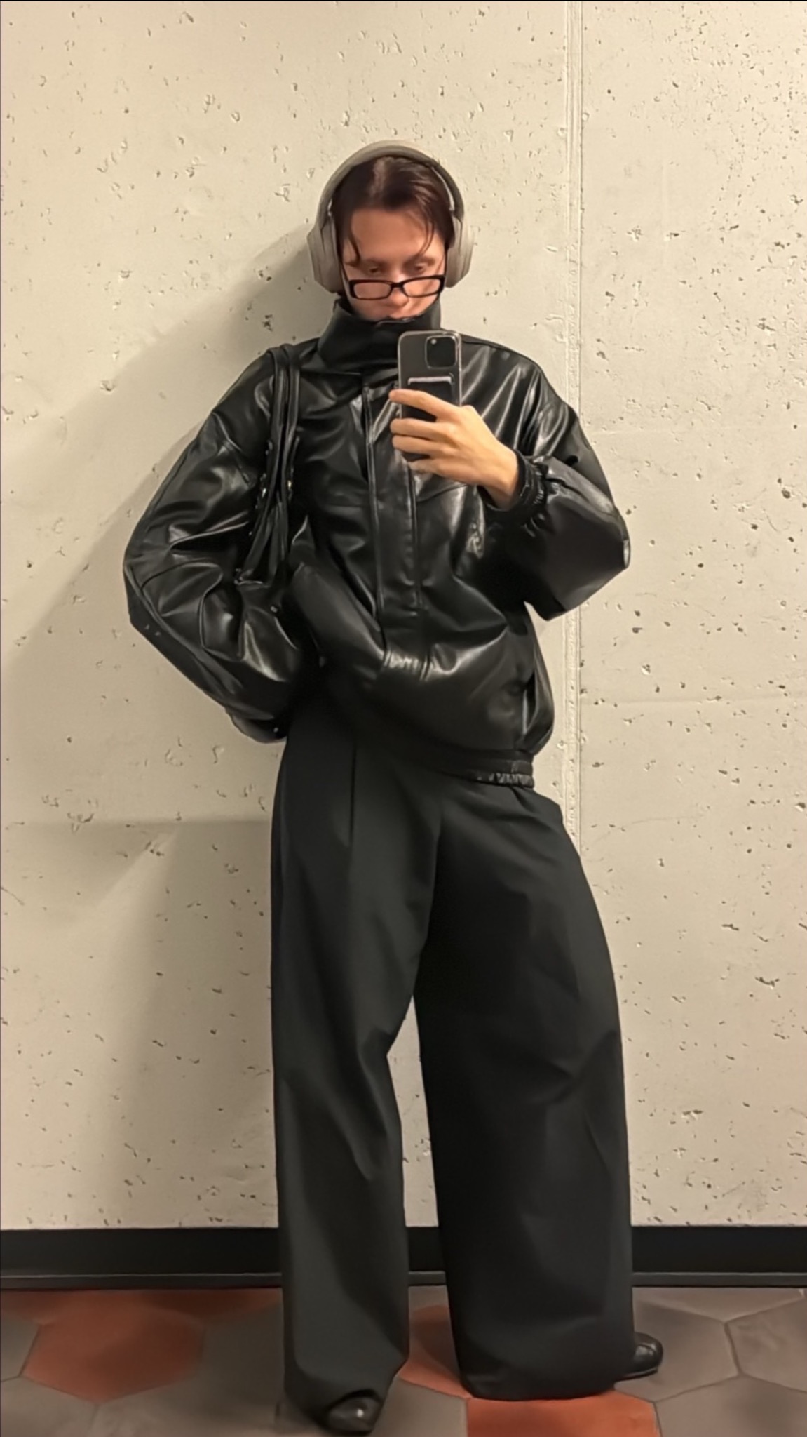 Osoba w czarnym, oversize'owym stroju pozuje do selfie. Widoczne słuchawki na uszach, okulary i torebka na ramieniu. Tło stanowi betonowa ściana.